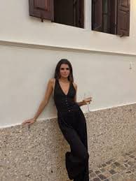 Mujer con mono negro y copa de vino apoyada en una pared blanca con contraventanas.